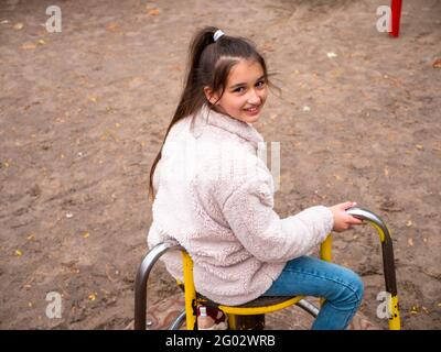 Top view over a cheerful smiling teenage girl (11-12) in a faux fur coat and with long ponytail spinning at a merry-go-round at playground. Stock Photo