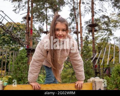 Happy smiling teenager girl in a faux fur coat and with long ponytail climbing up the yellow climber at children playground. Rope park with pine trees Stock Photo