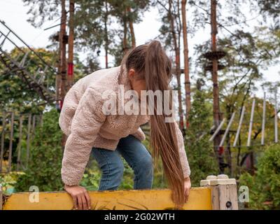 Happy smiling teenager girl in a faux fur coat and with long ponytail climbing up the yellow climber at children playground. Rope park with pine trees Stock Photo