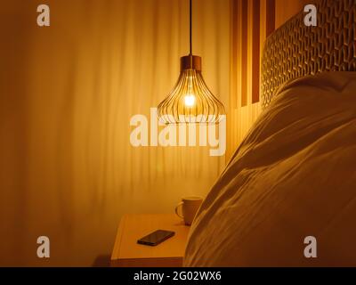 Metal lamp shade with yellow light above the bedside cabinet with smartphone and a cup on the background and blurry big pillow on the foreground. Stock Photo