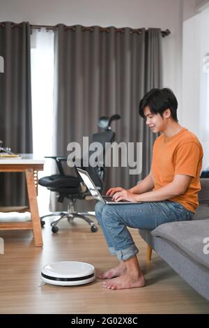 Casual man using laptop on sofa and robotic vacuum cleaner cleaning in living room. Stock Photo