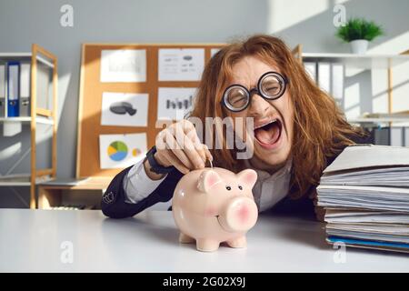 Happy crazy looking young man making funny face and putting coin in pink piggy bank Stock Photo