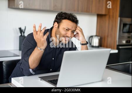 Confused indian man in smart casual shirt looking at the laptop screen, misunderstanding what happened, issues with a project or system error on the computer Stock Photo