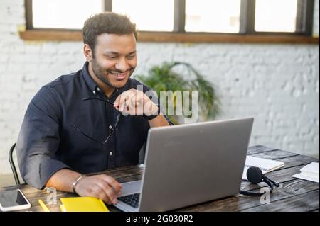 Optimistic indian mixed race man using laptop, male employee in smart casual shirt take off eyewear, watching at the monitor and smiling Stock Photo