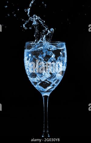 A wineglass with ice cubes and splashes, on a black background, toned in a blue tint. Stock Photo