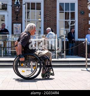 Epsom Surrey London UK, May 31 2021, Senior Disabled Man In A Wheelchair Moving Along A Pedestrianised Area With People Drinking In The Backgorund Stock Photo