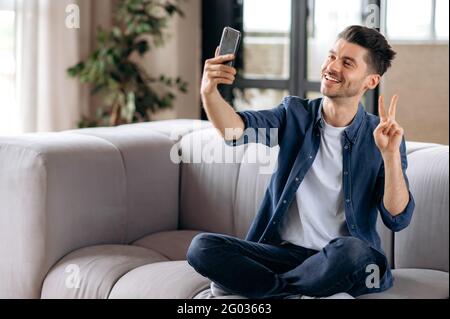 Happy cheerful handsome caucasian man, in casual clothes, resting, sitting on the sofa in the lotus position, holding a mobile phone in his hand, taking a selfie, gesturing with his hand, smiling Stock Photo
