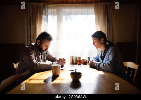 Portrait of sad poor mature couple praying at the table indoors at home, poverty concept. Stock Photo