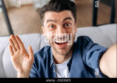 Webcam headshot of joyful handsome cheerful young caucasian man, in stylish casual clothes, sitting on the couch, uses smart phone, taking a selfie, looks at webcam, gesturing with his hand, smiling Stock Photo