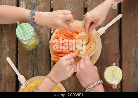 Group of unrecognizable happy friends taking hummus with carrot sticks in a park having picnic on a sunny summer day. Top view. Stock Photo