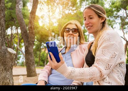 Happy mother and her daughter looking smartphone making a video call or watching a video online at sunset in nature park. Stock Photo
