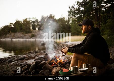 Side view of thoughtful man looking away while sitting by campfire at lakeshore Stock Photo