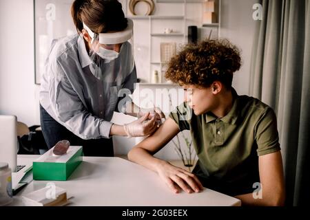 Female nurse giving COVID-19 vaccine to teenage boy at home Stock Photo