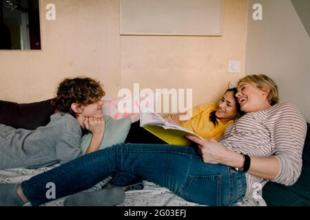 Smiling boy looking at cheerful mother and sister with book while lying on bed Stock Photo