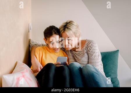 Mature woman sitting with daughter using smart phone in bedroom at home Stock Photo