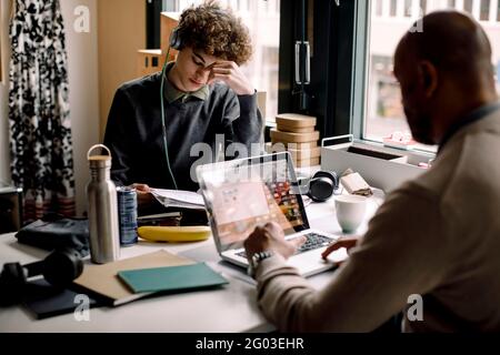 Boy wearing headphones studying while sitting with businessman using laptop at desk Stock Photo