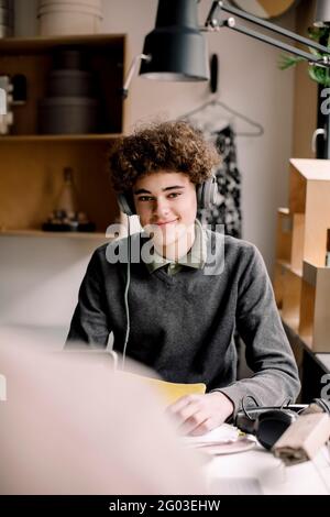 Portrait of smiling curly hair boy wearing headphones sitting at desk Stock Photo
