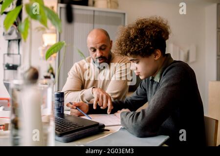 Father guiding son doing homework while sitting at table Stock Photo