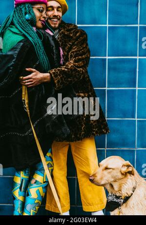 Smiling young man hugging woman while standing with dog against blue tiled wall Stock Photo