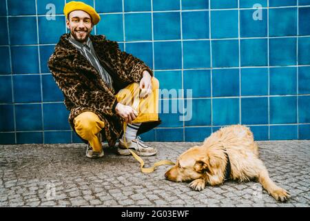 Full length of young man crouching by dog lying on footpath against blue tiled wall Stock Photo