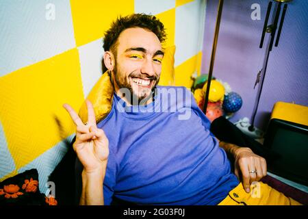 Portrait of happy young man gesturing peace sign while sitting in living room Stock Photo