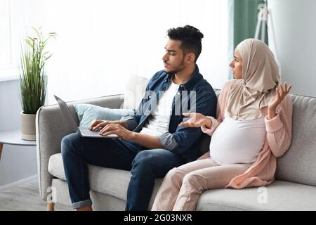 Annoyed pregnant muslim woman looking at husband that spending time with laptop Stock Photo