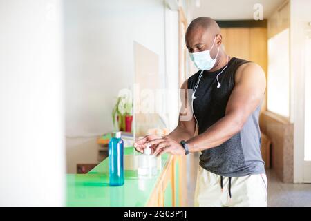 Black man in sportswear and face mask spraying sanitizing gel on his hands. He is entering the gym. Stock Photo