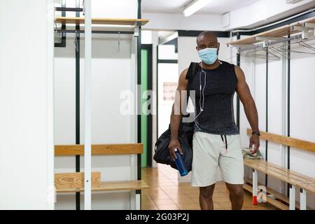 Young black male in sports clothes in gym locker room. He wears a medical mask as a protective measure against the Covid-19 coronavirus. Stock Photo