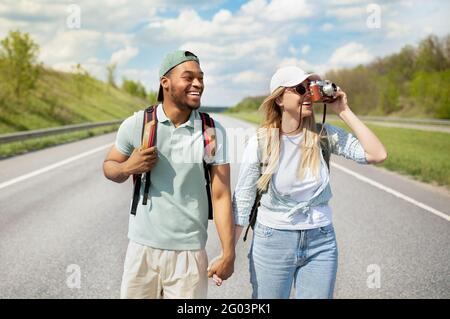 Happy young multiracial couple hitchhiking together, holding hands, taking photos on camera, walking along road Stock Photo