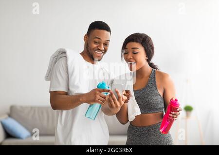 Home workout. Excited black couple holding bottles of water, using cellphone, ready for domestic training, indoors Stock Photo