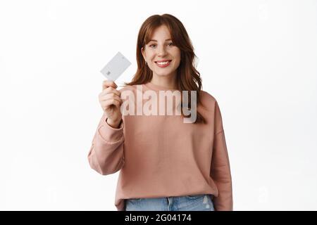 Portrait of redhead girl smiling, showing credit card, advertising bank, special offers or discounts, going on shopping, standing over white Stock Photo