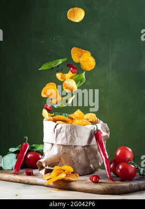 Potato chips fall into a paper bag. Spicy chips in a craft bag. Flying potatoes and spices. Flying chips. Vertical frame. Stock Photo
