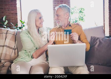 Portrait of beautiful elderly retired pensioner cheerful couple sitting on sofa using card buying web store at home brick loft interior house indoor Stock Photo