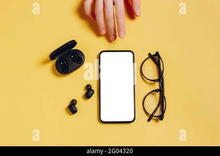 Hand reaching for smartphone with blank empty white screen. Mobile app mockup with wireless headphones, charger, and glasses on yellow background Stock Photo