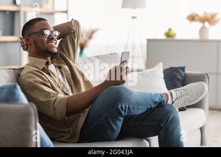 Cheerful Black Man Using Cellphone Wearing Eyeglasses At Home, Side-View Stock Photo