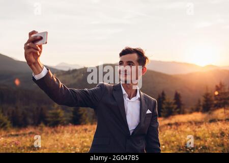 Handsome groom taking selfie in wedding suit in mountains at sunset. Smiling man holding smartphone Stock Photo