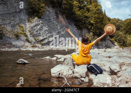 Hiker woman relaxing by mountain river enjoying landscape. Traveler raising arms sitting on rocks. Summer vacation Stock Photo