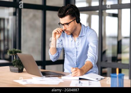 Concentrated caucasian man with glasses and headphones, office worker or consultant, stands near workplace in the office, communicates with colleagues or client on an online conference, takes notes Stock Photo