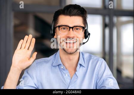 Portrait friendly joyful young adult caucasian business leader, manager or consultant wearing glasses and headset, communicating via video conference with employees or client, waving hand, smiling Stock Photo