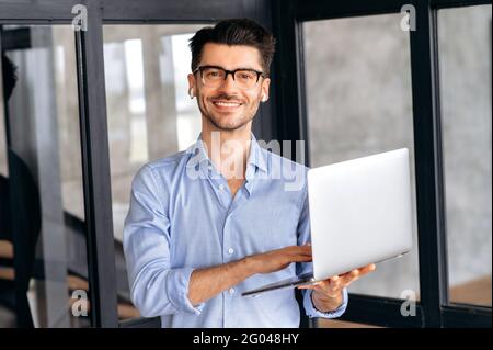 Portrait of young adult successful intelligent caucasian businessman, or ceo, wearing glasses and formal stylish clothes, standing in office, holding open laptop in his hand, looks at camera, smiling Stock Photo
