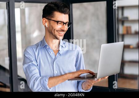 Joyful successful caucasian businessman, manager or ceo, wearing glasses, standing in office, holding and uses laptop, chatting online with colleagues or client, browsing internet, smiling Stock Photo