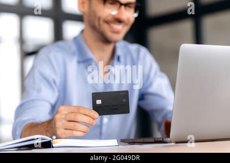 Blurred joyful successful caucasian man wearing glasses, in formal clothes sitting at table, using laptop and credit card to pay for goods and delivery via Internet, shopping online Stock Photo