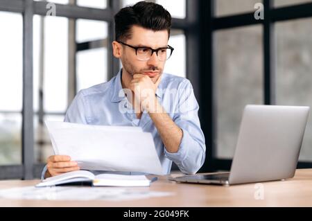Successful smart focused young caucasian business man, freelancer or manager sitting at the table in the office, looking through documents, thinking over business strategy, looking at laptop screen Stock Photo