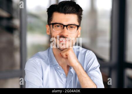 Close-up portrait of a young handsome caucasian guy in glasses, in stylish formal clothes, sitting in the office. Confident intelligent male business person looks at camera, smiles friendly Stock Photo