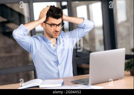 Upset angry shocked caucasian businessman, broker or ceo, wearing glasses, working in office, using laptop, overworked, experiencing stress holding hands behind his head, irritated executive Stock Photo