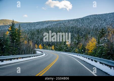 Autumn maples and winter snow-covered winding road with first snow dusted trees on the mountains in Vermont invite tourists and travelers to visit the Stock Photo