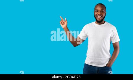 Black Man Pointing Finger Aside Smiling To Camera, Blue Background Stock Photo