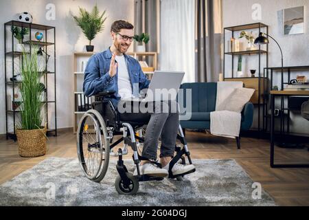 Bearded young man sitting in wheelchair with portable laptop and talking during video call. Male freelancer with special needs having online working meeting. Stock Photo