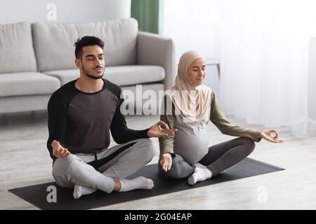 Pregnant Muslim Woman In Hijab Practicing Yoga With Her Husband At Home Stock Photo