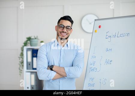 Distance learning. Happy Arab male teacher giving online English class, standing with crossed arms near blackboard Stock Photo